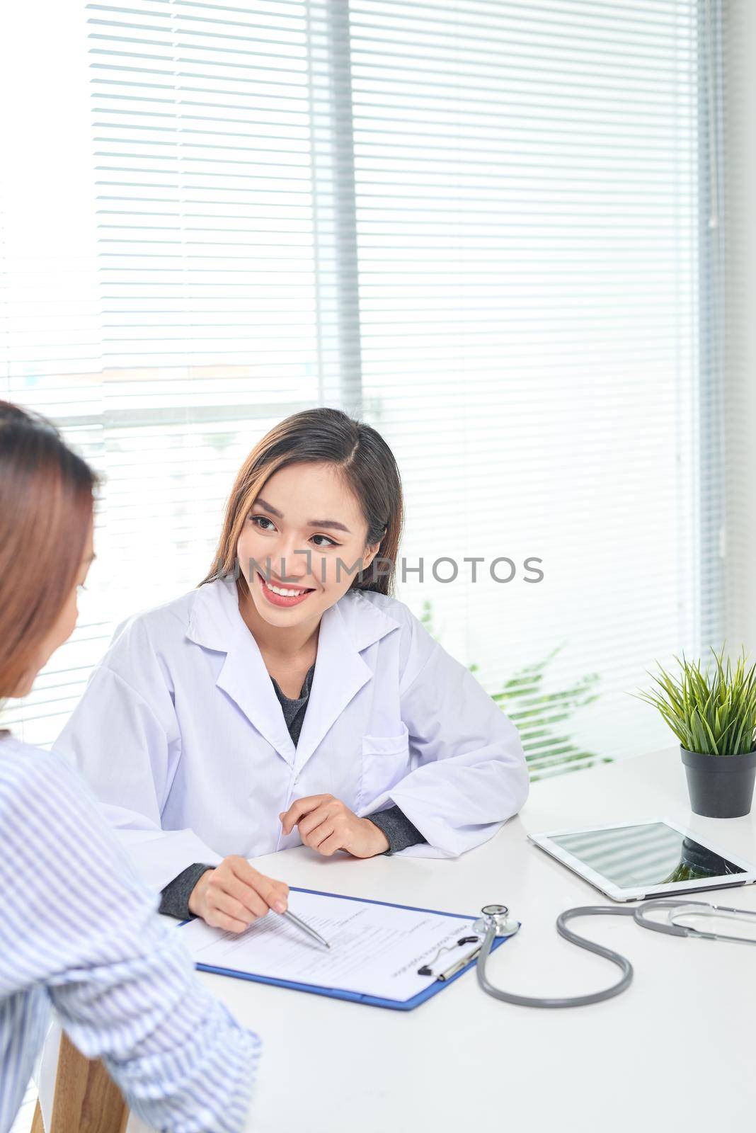 Female doctor talks to female patient in hospital office while writing on the patients health record on the table. Healthcare and medical service.  by makidotvn