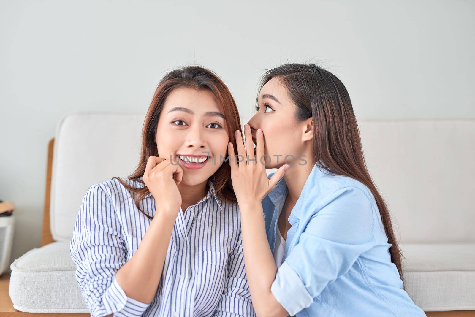 portrait of two young attractive happy women with  eavesdrops whispers a secret (mystery) in the studio on living room. The concept of gossip and confidentiality by makidotvn