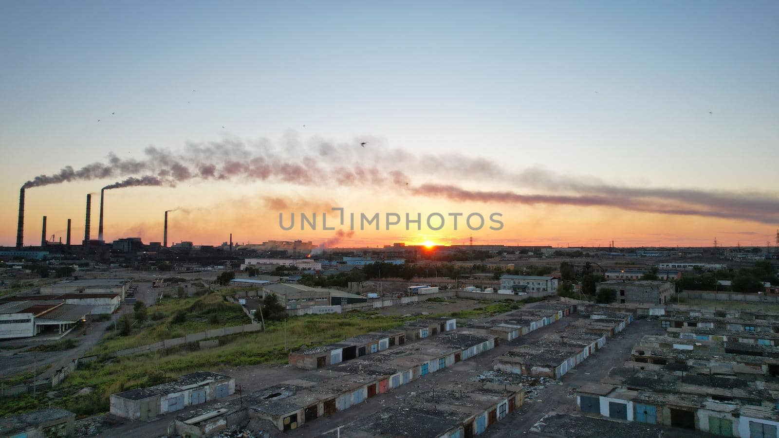 An epic sunset with a view of the smoking factory and the city. The embankment of Lake Balkhash. Low houses are standing and there are few trees. The pipes from the factory are smoking. Kazakhstan