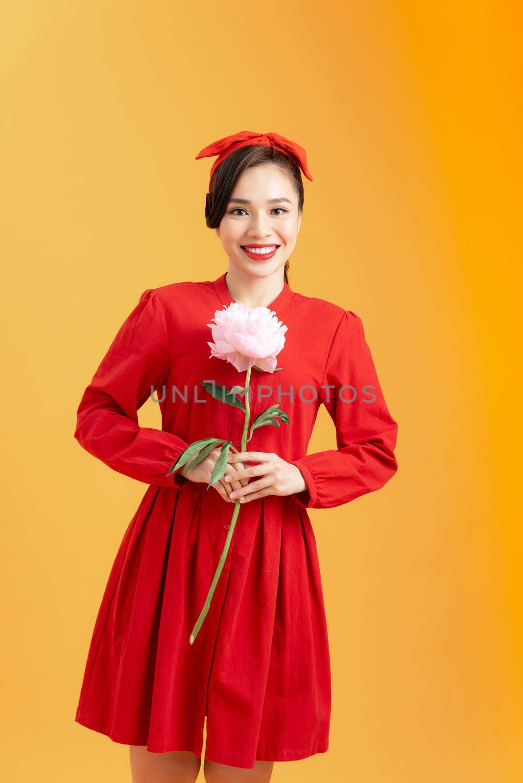 Portrait of an attractive young Asian woman in red dress holding peony flower isolated over orange background