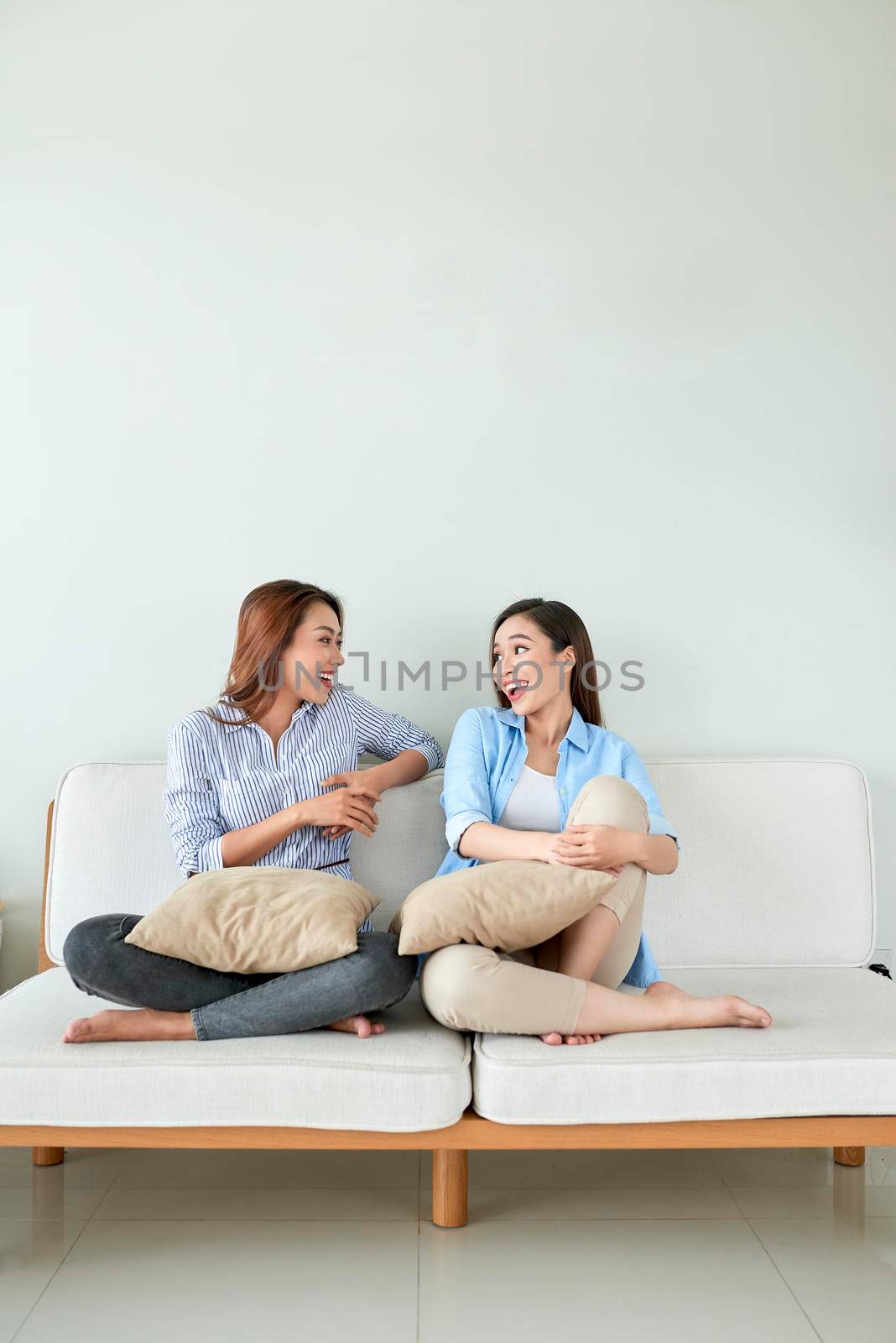 Two young beautiful cheerful girlfriends sitting on sofa at home laughing.