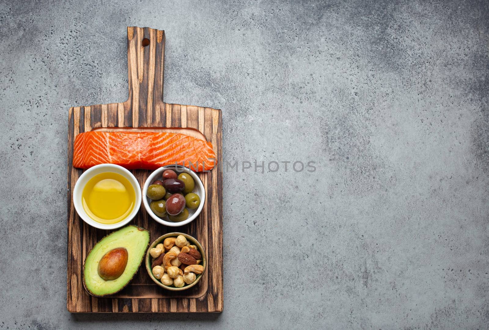 Food sources of healthy unsaturated fat and omega 3: fresh raw salmon fillet, avocado, olives, nuts on cutting board, rustic stone background top view. Healthy nutrition and keto diet, space for text