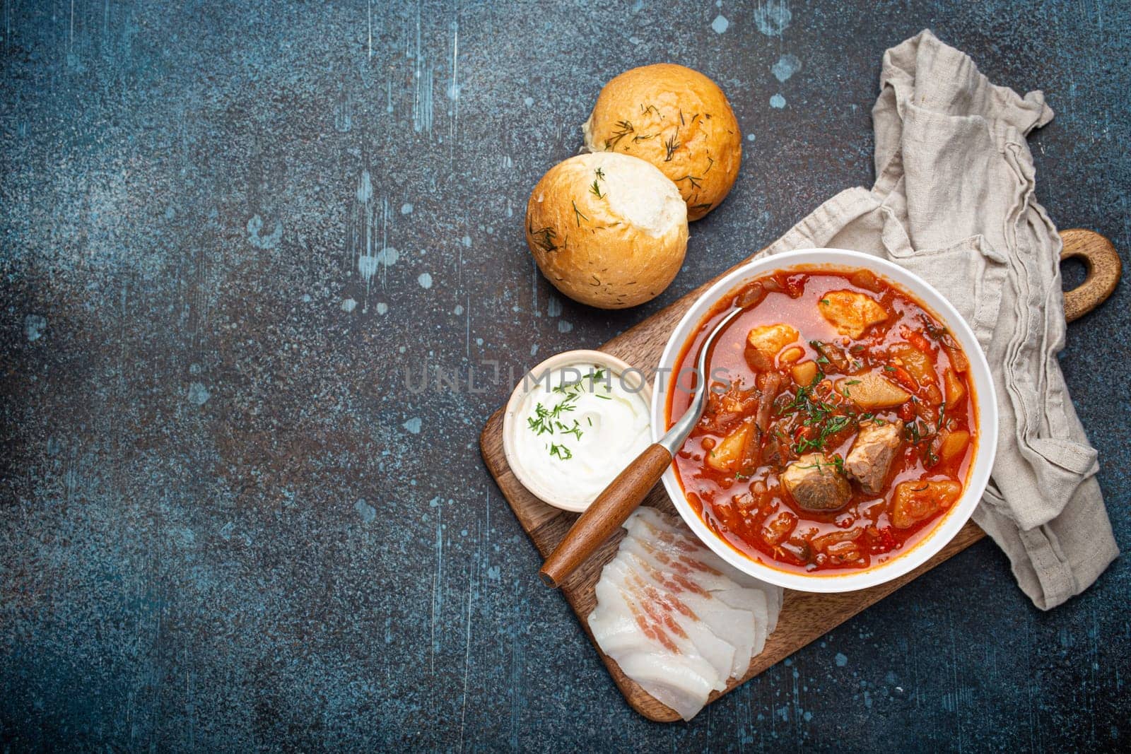 Ukrainian Borscht, red beetroot soup with meat, in white bowl with sour cream, garlic buns Pampushka and salo slices on rustic stone background. Traditional authentic dish of Ukraine, space for text