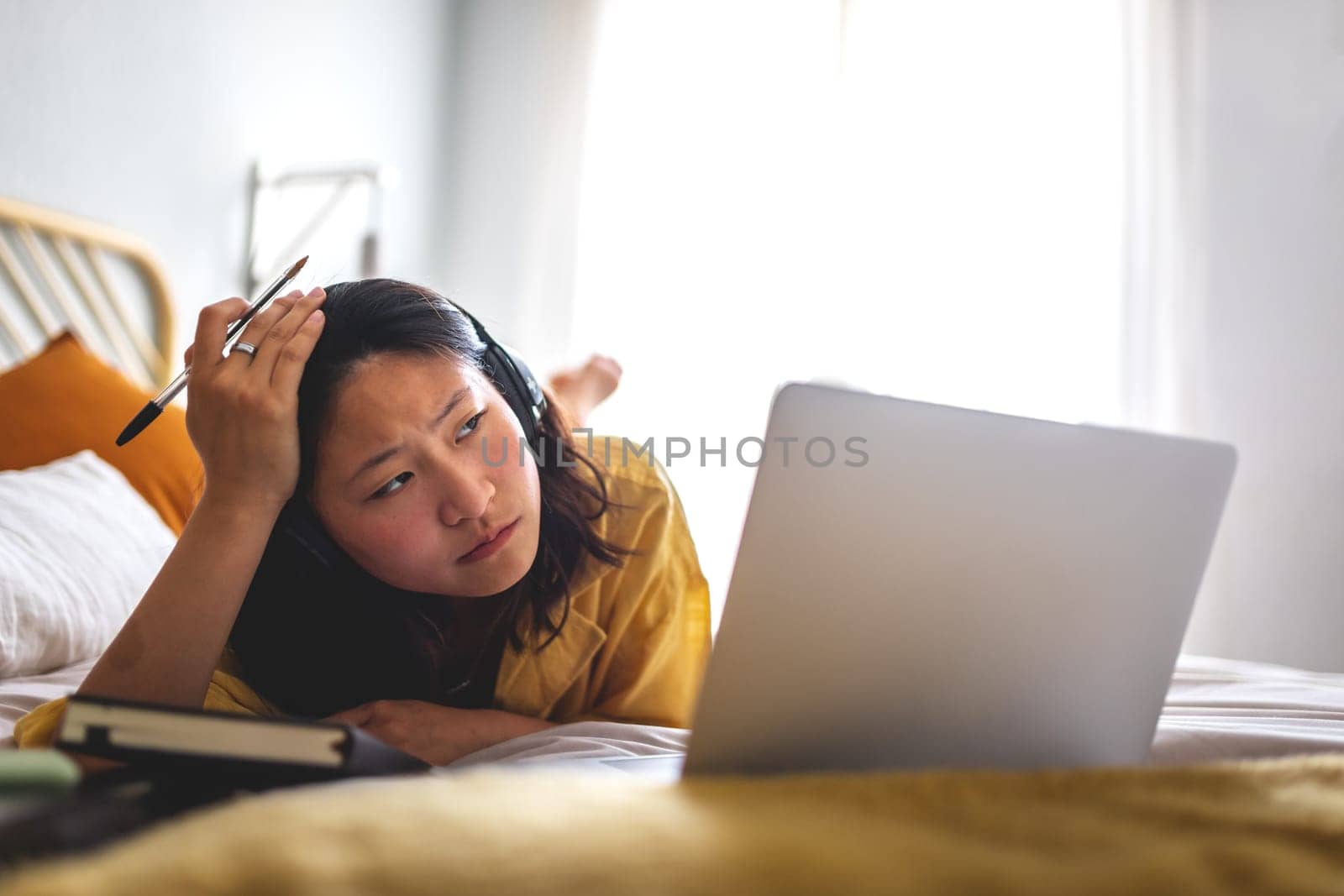 Stressed, worried Asian teen girl on bed attending online classes listening teacher lesson with headphones using laptop by Hoverstock