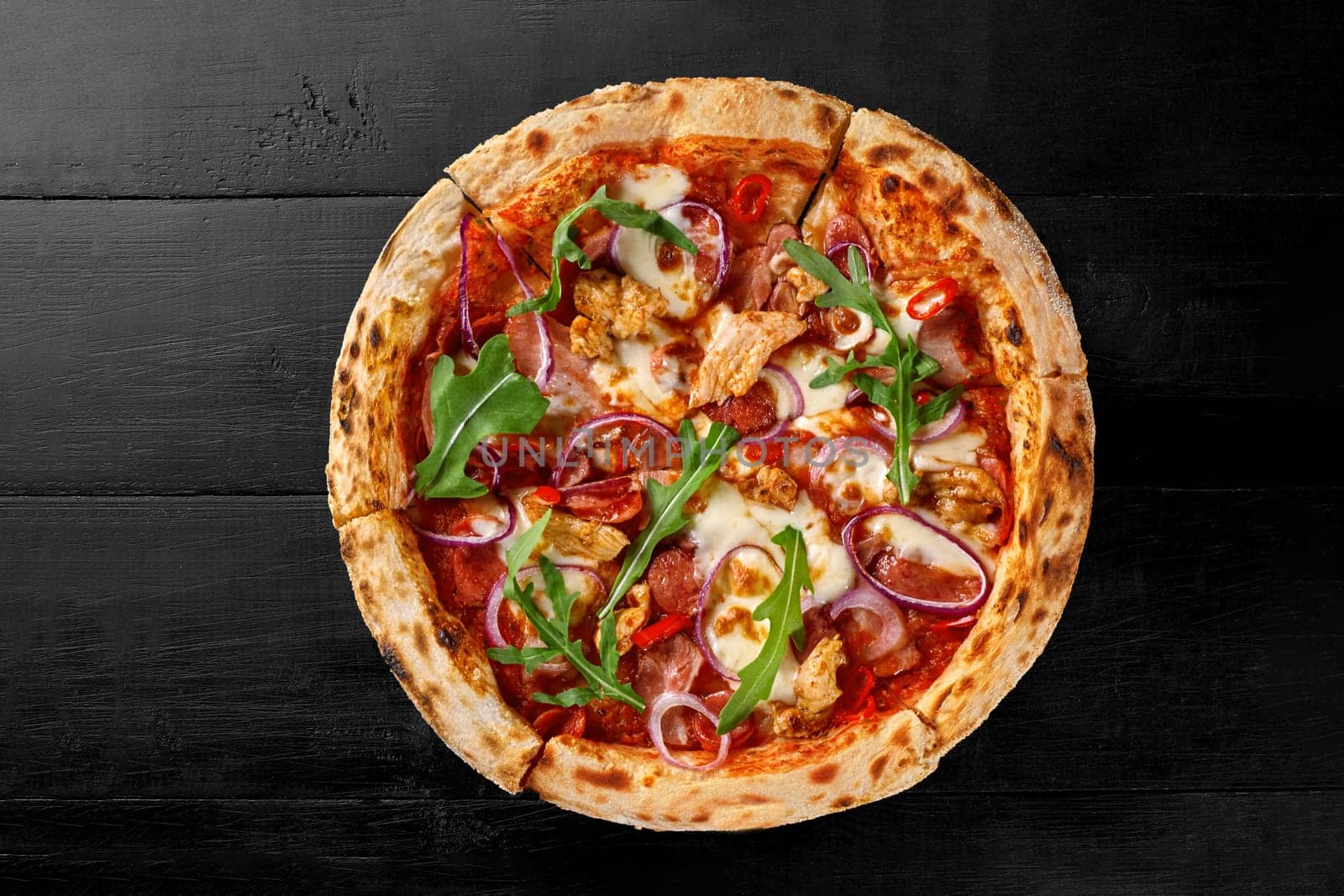 Appetizing browned pizza with chicken fillet, bacon, salami, hunting sausages, chili pepper, onion rings and greens on pelati sauce and melted mozzarella on black wooden background. Italian cuisine