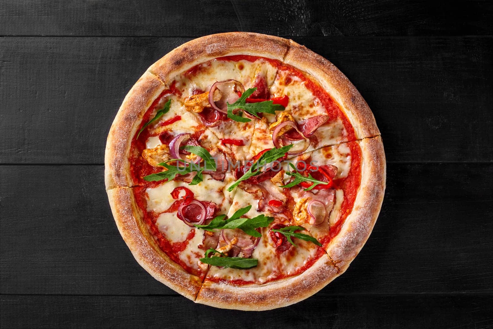 Delicious pizza with browned edge filled with sous vide chicken fillet, bacon, salami sausages, spicy pepper, purple onion rings and arugula on black wooden background