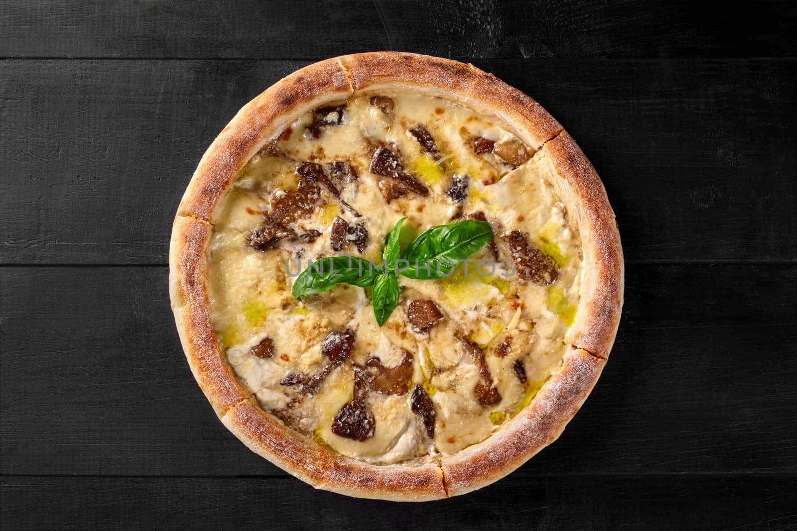 Delicious pizza with delicate creamy cheese sauce, fried forest mushrooms and champignons garnished with fresh fragrant basil on black wooden surface. Popular Italian style food