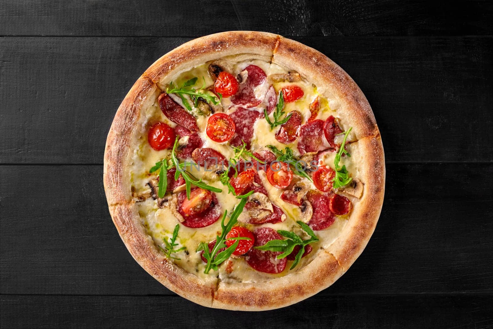 Delicious freshly baked Italian Boscaiola pizza with cream sauce, mozzarella, slices of cabanossi and salami, mushrooms, parmesan garnished with fresh arugula leaves on black wooden surface
