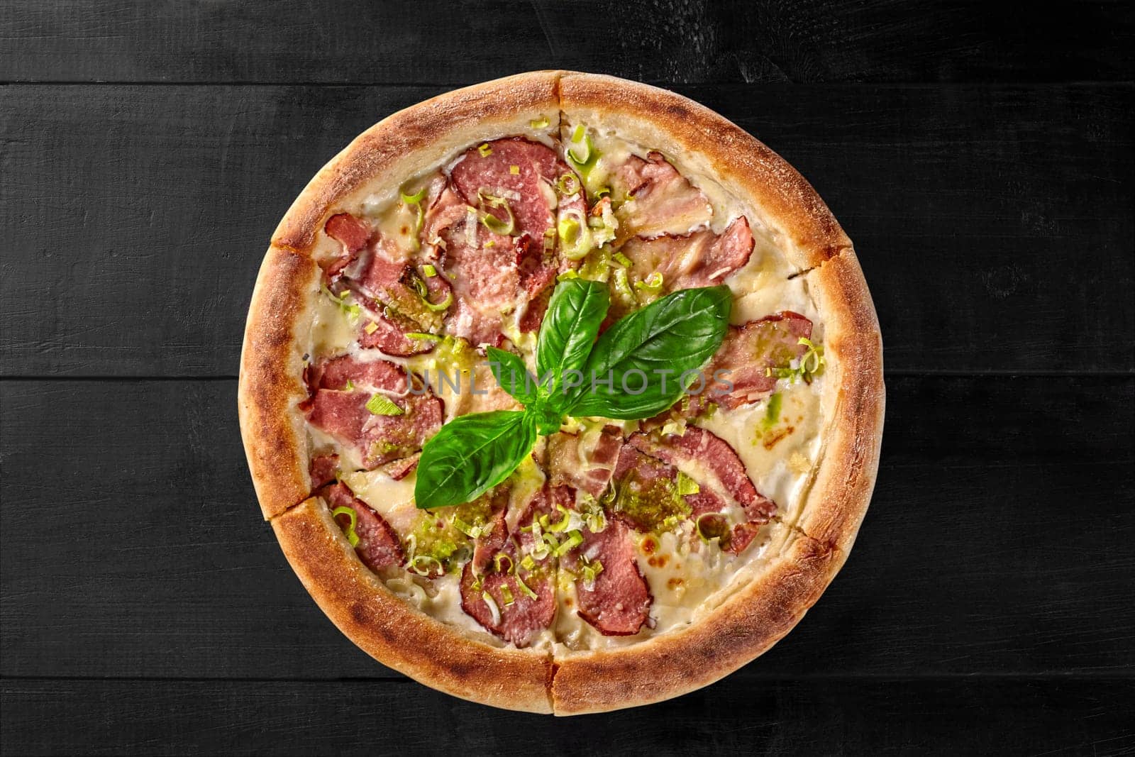 Top view of appetizing pizza with cream cheese sauce topped with smoked bacon and ham slices seasoned with pesto sauce garnished with fresh basil and shallot on black wooden background