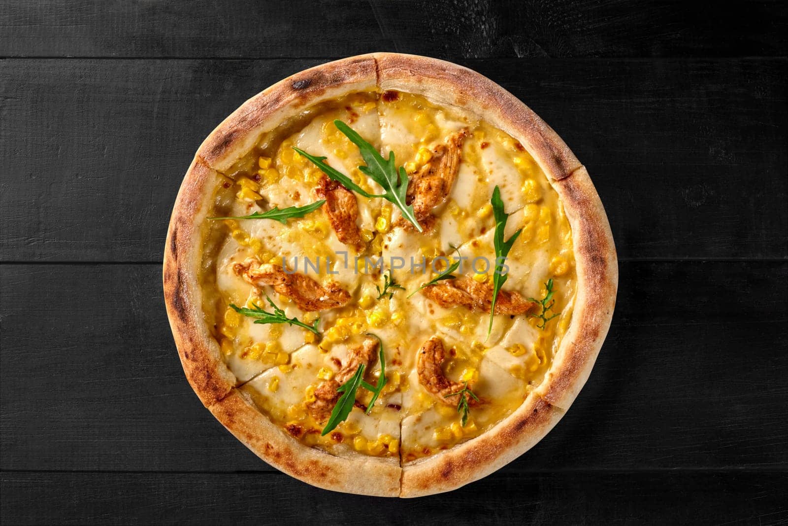 Top view of delicious savory pizza with melted cheese, corn kernels and sous vide chicken filet topped with green arugula on black wooden background