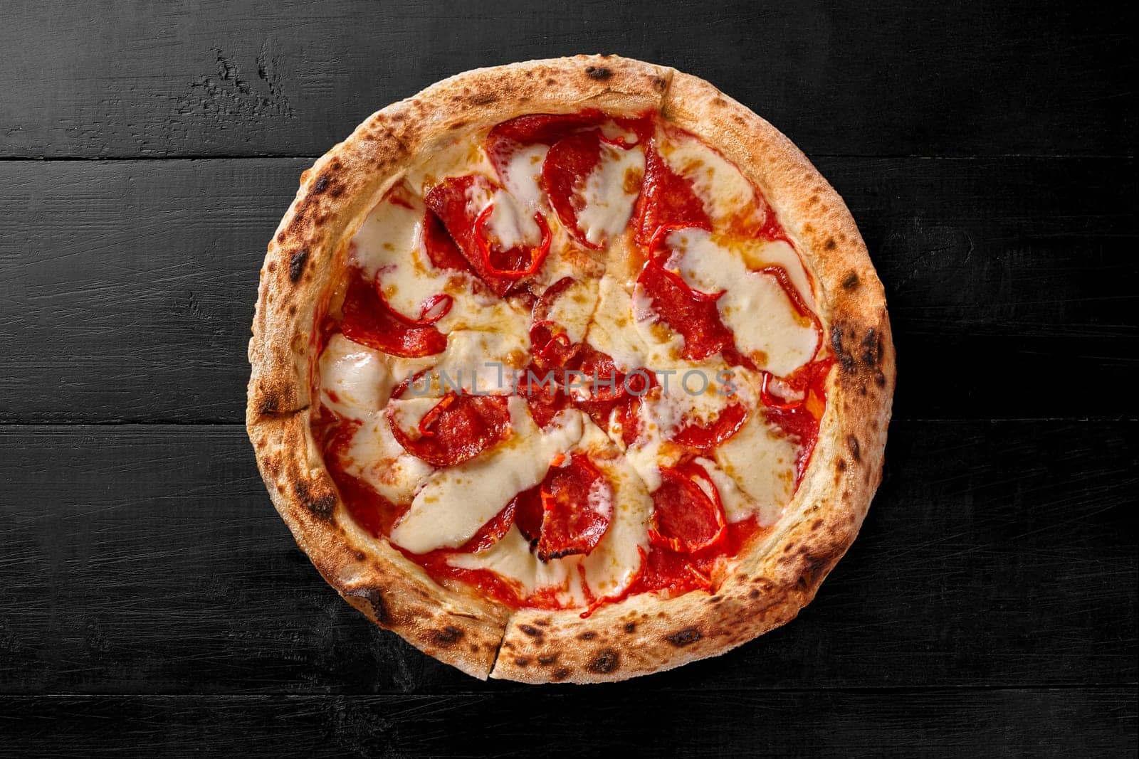 Delicious pepperoni pizza with browned dough edge, mozzarella cheese, thinly sliced salami sausage and spicy red chili pepper on black background. Italian-American cuisine