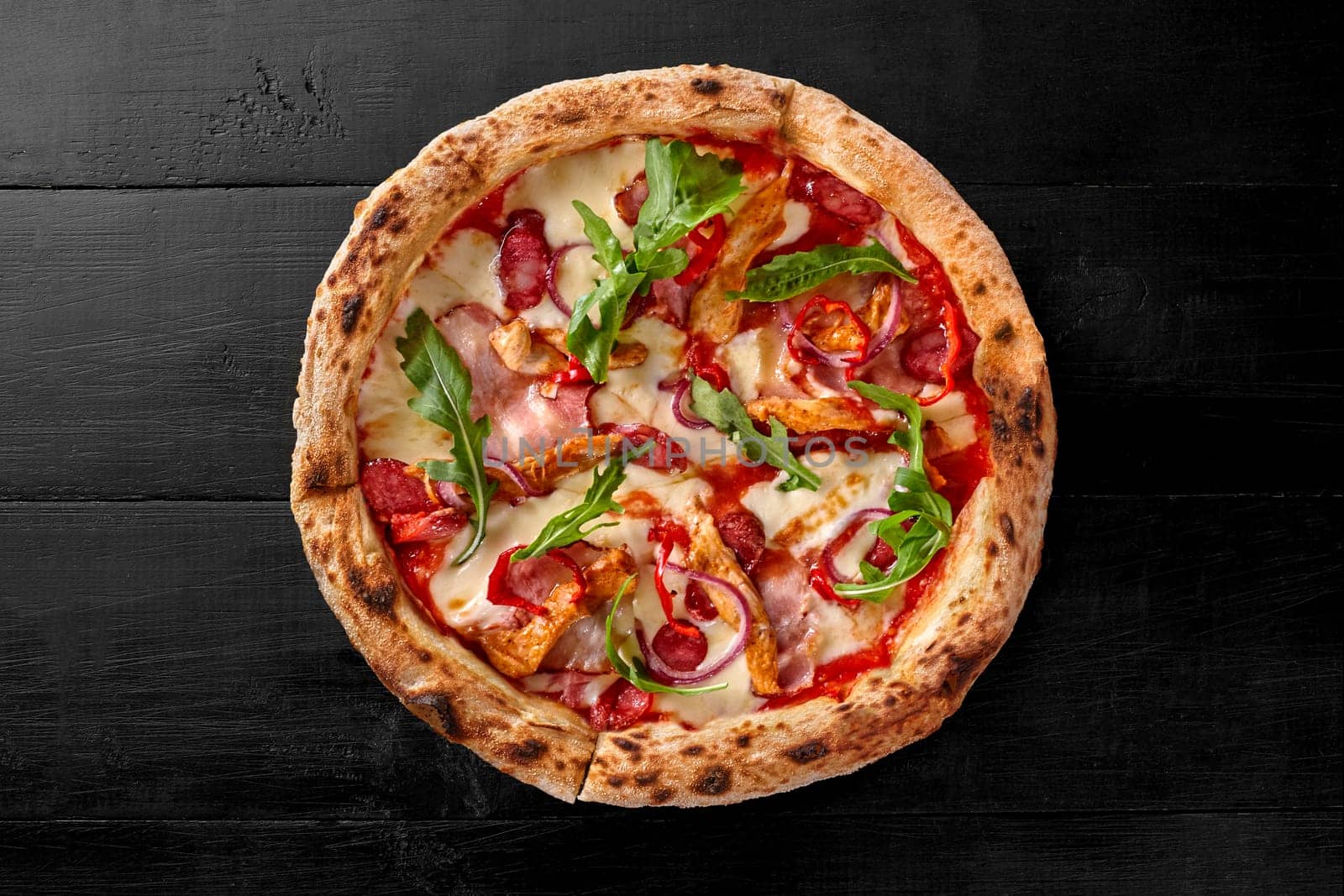 Appetizing thin pizza with browned edge filled with pelati sauce, cheese, hunting sausages, bacon slices, sous vide chicken, rings of onion and chili pepper and green arugula on black wooden surface