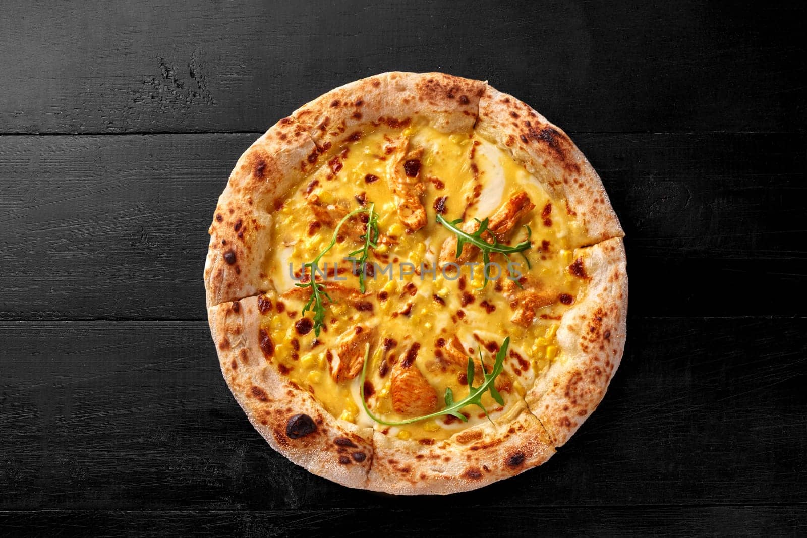 Delicious sous vide chicken pizza on thin dough with corn, browned edge and melted cheese base garnished with fresh green fragrant arugula on black wooden surface, top view