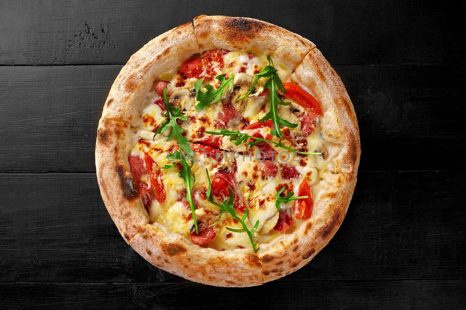 Delicious freshly baked pizza with cream cheese sauce, salami and bacon slices, mushrooms and tomatoes garnished with fresh fragrant arugula served on black wooden background