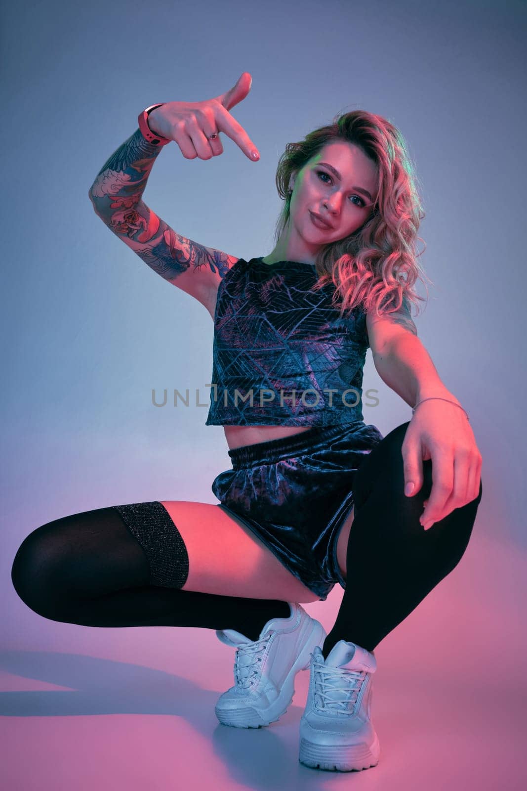 Beautiful young blonde tattoed girl in blue velour booty shorts and t-shirt, black stockings, squats in front of the camera in a confident pose. Pink and blue background