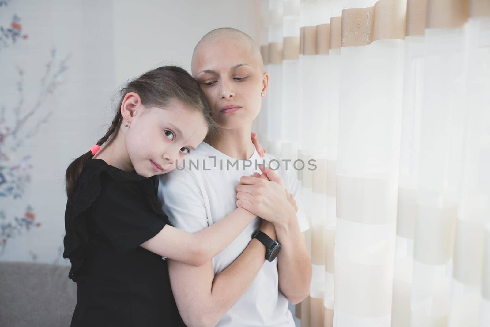 Kid girl hugs her mother with a diagnosis of leukemia near the window. Family support concept.