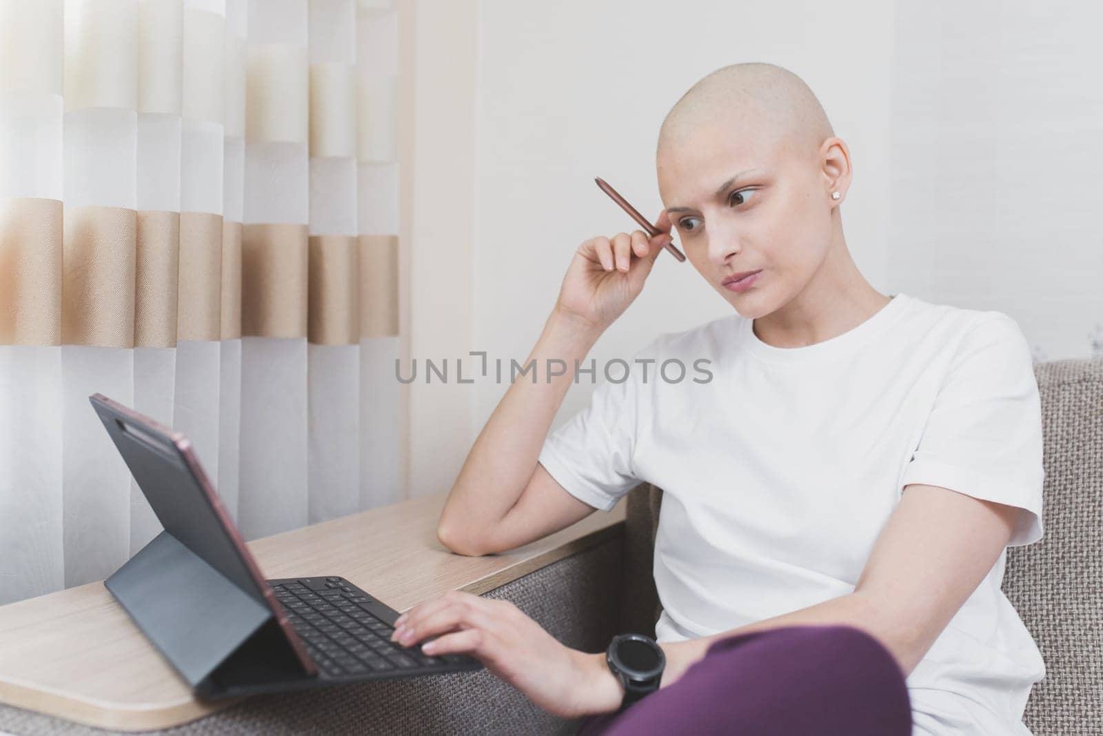 Hairless young woman with cancer wearing a white t-shirt working at laptop in room.