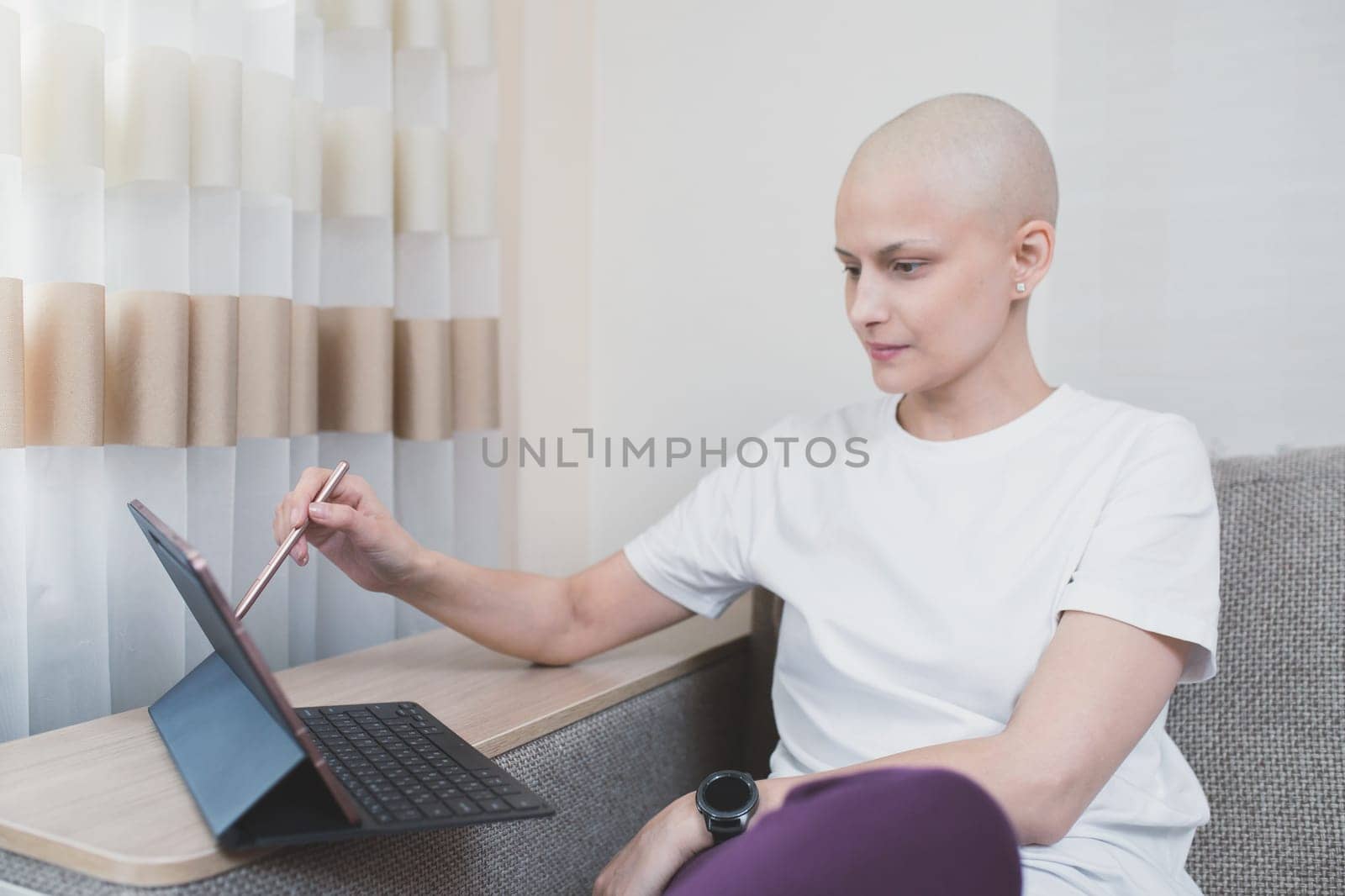 Young bald woman looks at laptop at home, distance learning or remote work, sick leave.