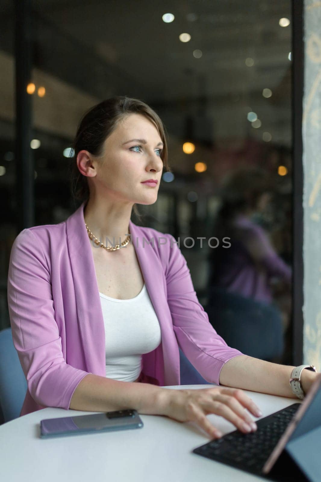 Portrait of a young beautiful business woman sitting in cafe with a laptop. Remote work in a coffee shop, look at window, vertical by Rom4ek