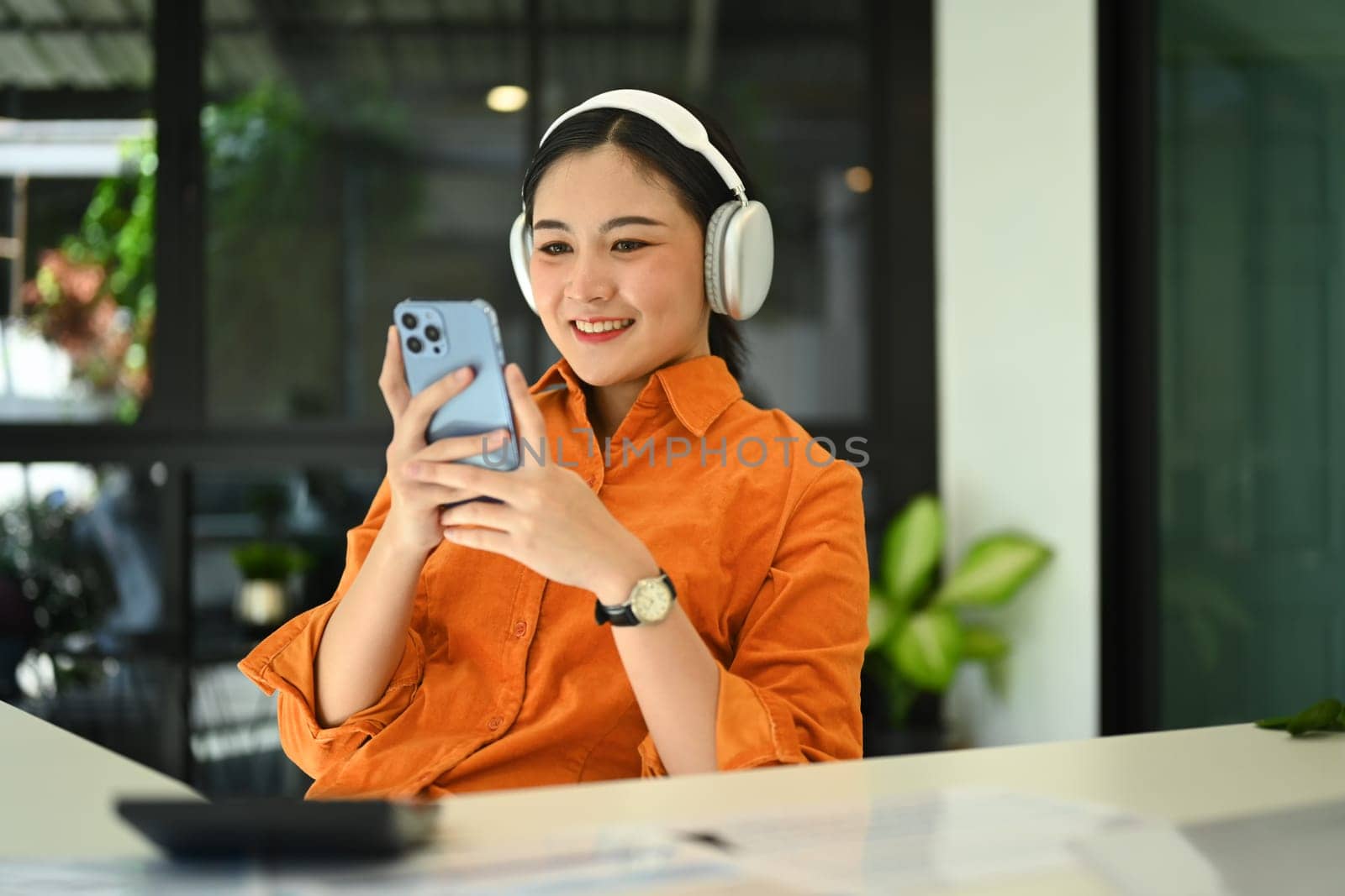 Cheerful stylish young woman wearing wireless headphone and using smartphone at her office desk by prathanchorruangsak