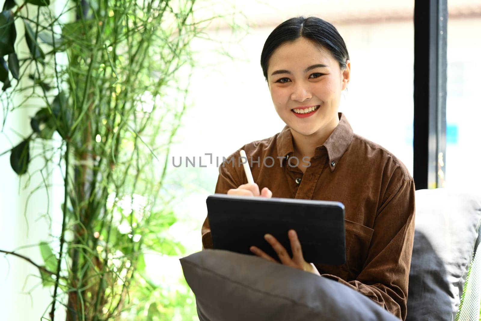 Pretty young office worker sitting on armchair in modern office and using digital tablet by prathanchorruangsak
