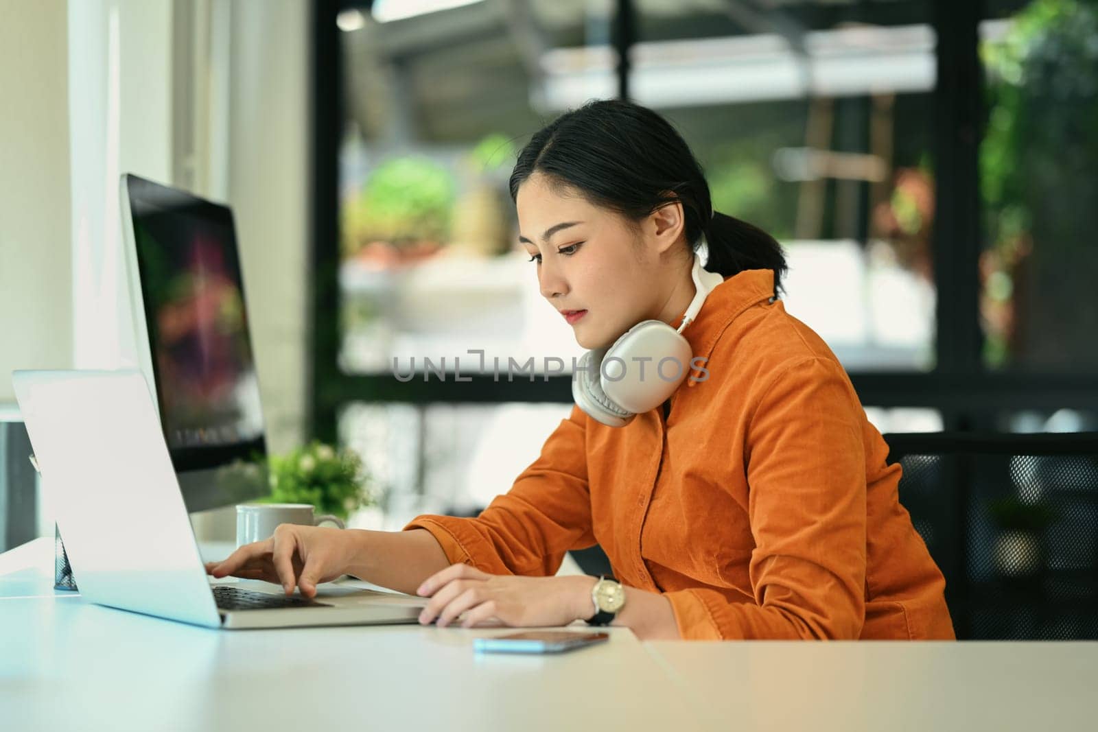 Focused young creative woman with headphone on her neck working on laptop at modern office.