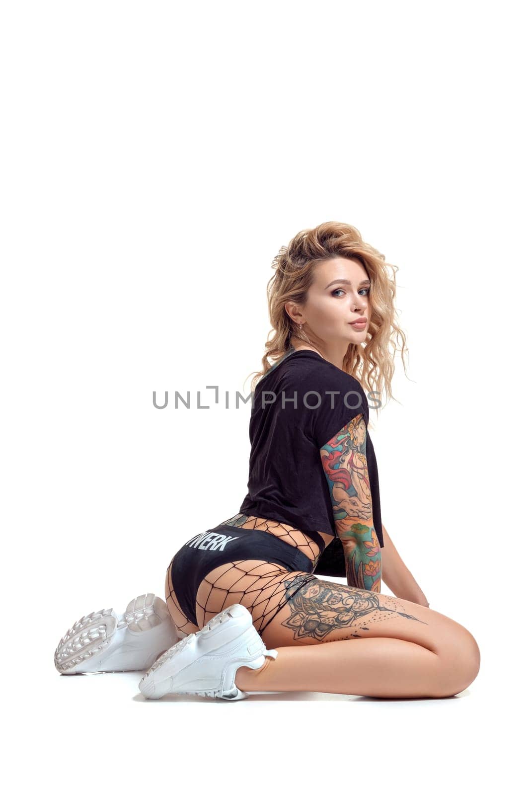 Wonderful blond twerk lady with tattoed body and long curly hair is posing sitting sideways on the floor and looking at the camera, isolated on white background with copy space. Young girl wearing in a mesh black shorts, top and white sneakers. Booty dance in studio.