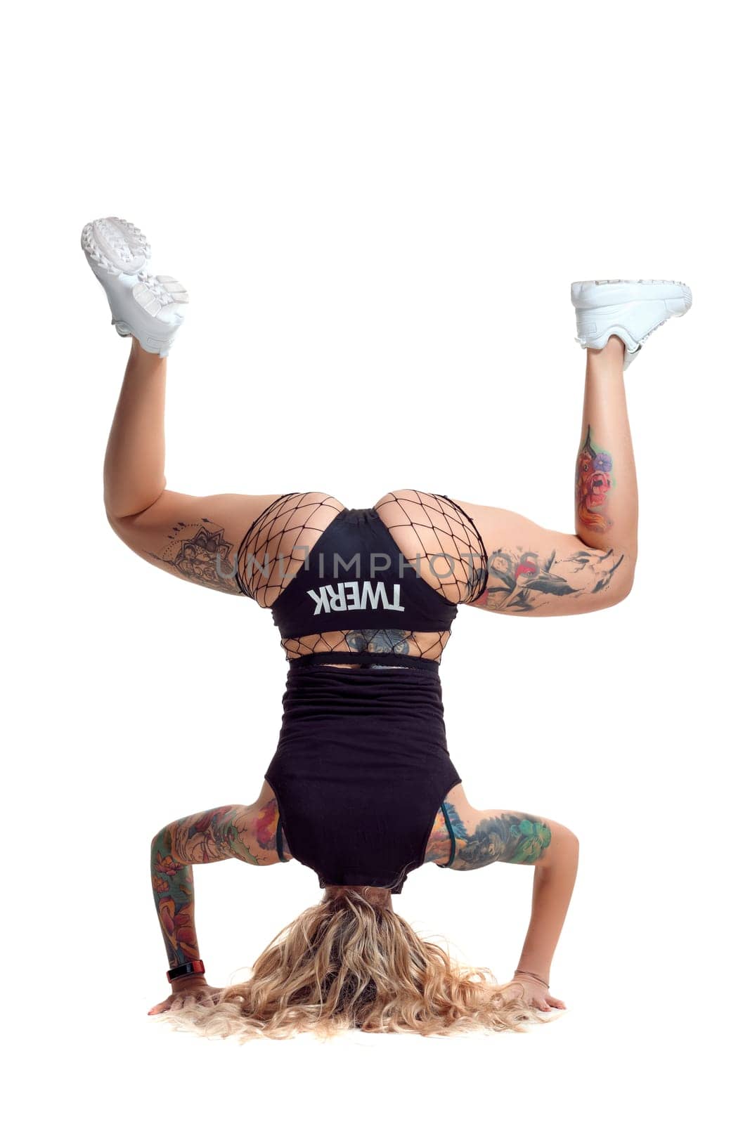 Nice blond twerk woman with tattoed body and long curly hair is posing standing on her head, isolated on white background with copy space. Attractive lady wearing in a mesh black shorts, top and white sneakers. Booty dance in studio.