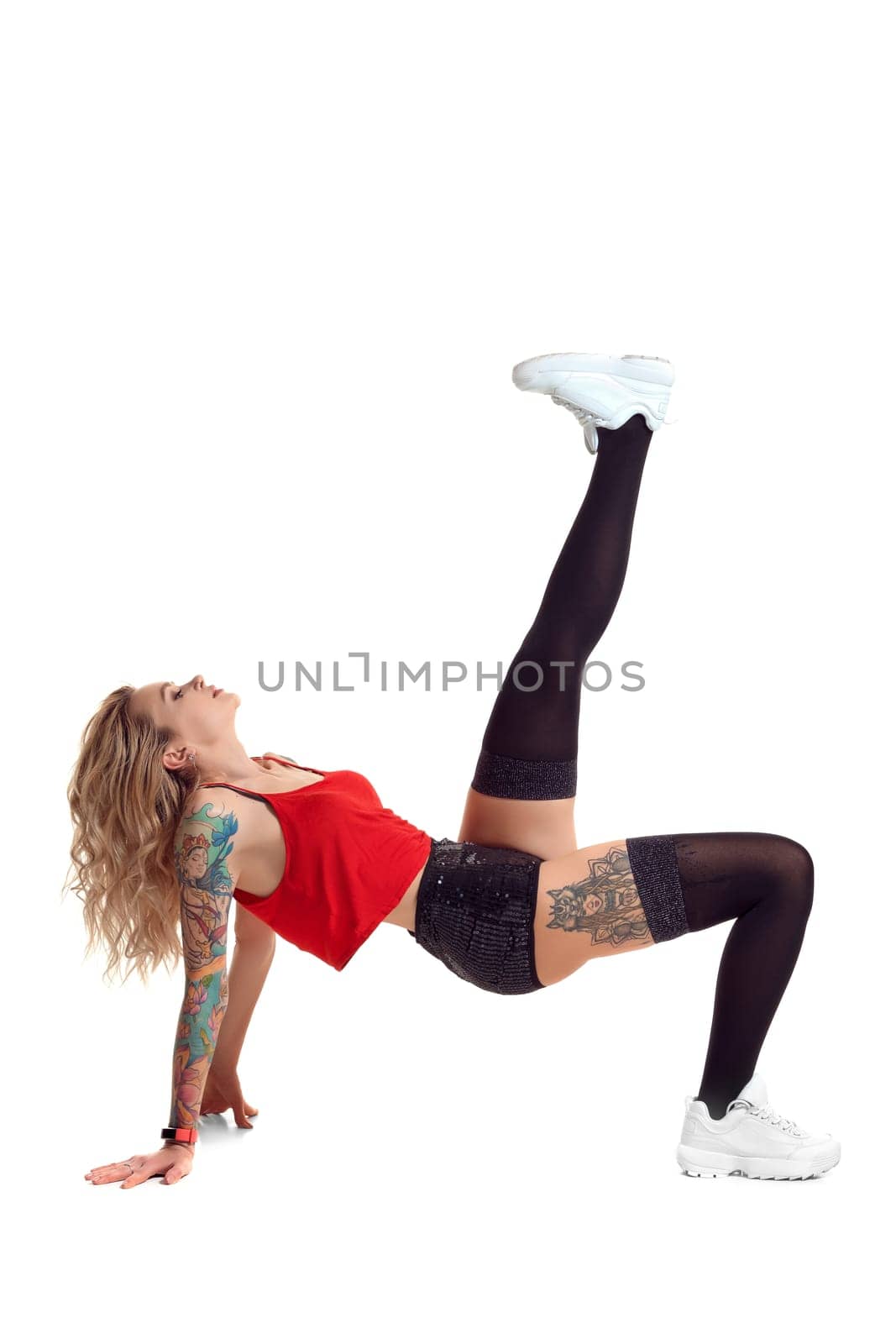 Gorgeous blond female with tattoed body and long curly hair is looking up while posing isolated on white background with copy space. Young woman wearing in a black stockings and mini shorts, red top and white sneakers. Booty twerk dance in studio.
