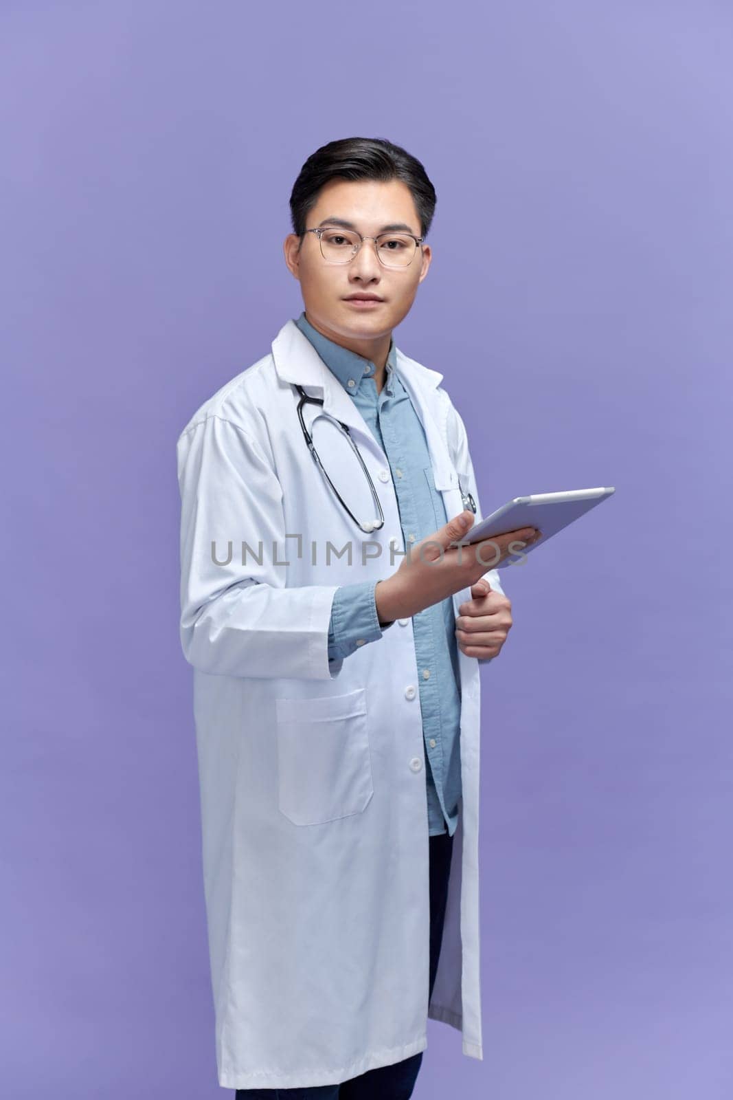smiling young male doctor in medical coat using digital tablet isolated on purple by makidotvn