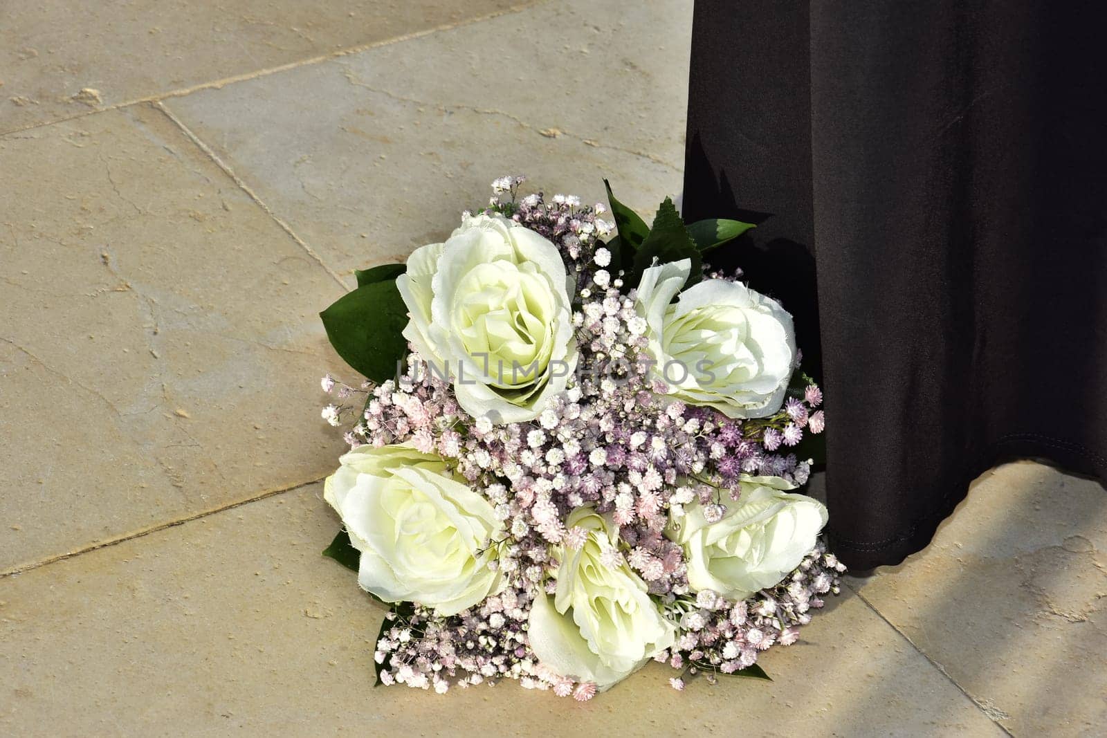 The bride's wedding bouquet of white roses and pink flowers lies on the marble yellow floor, next to the long black skirt of a standing woman. High quality photo