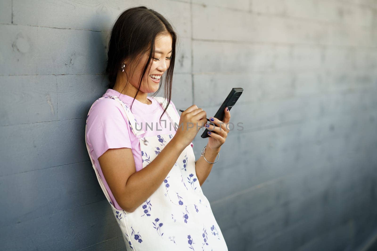 Happy Young Chinese woman using her smartphone with a pen or stylus, outdoors Asian female wearing casual clothes.