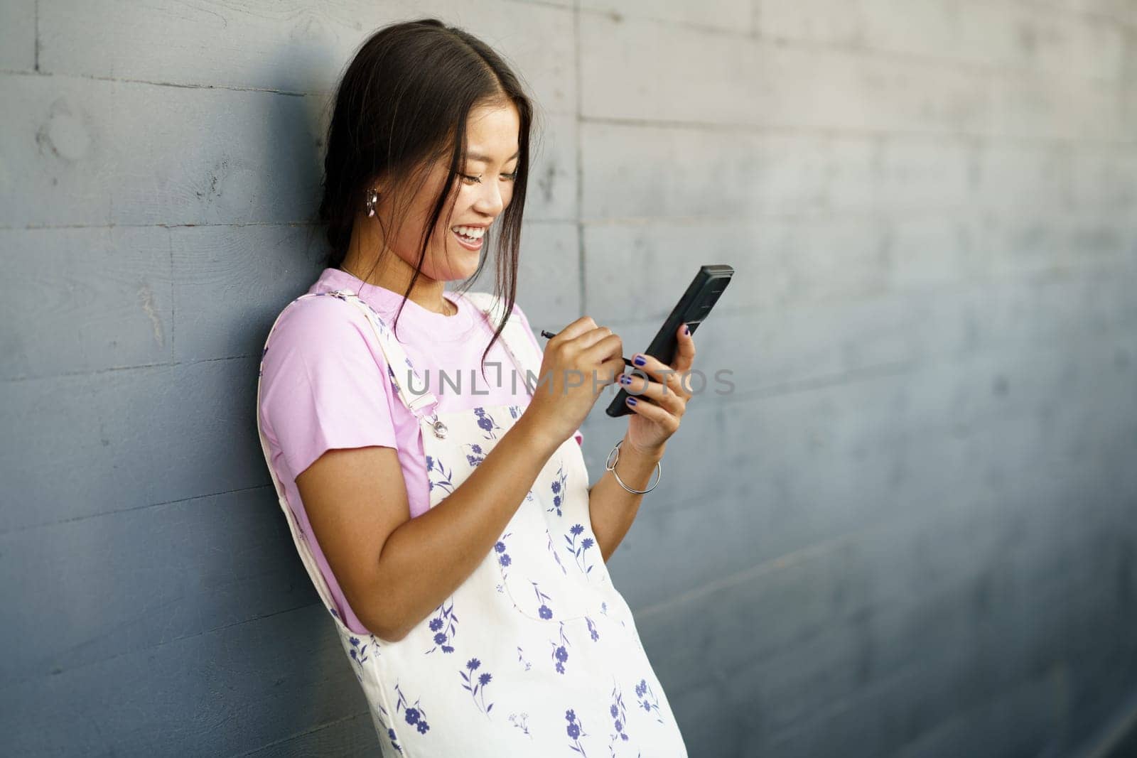 Happy Young Chinese woman using her smartphone with a pen or stylus, outdoors. by javiindy