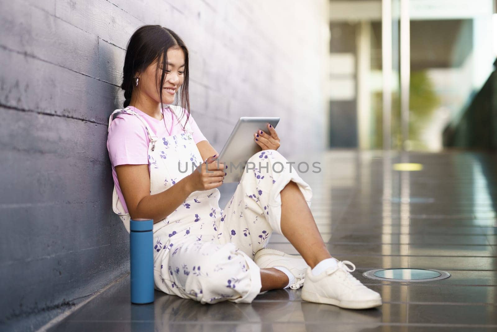 Full body glad Asian female in stylish outfit smiling and watching video on tablet while sitting on slope near thermos outside modern building in daytime