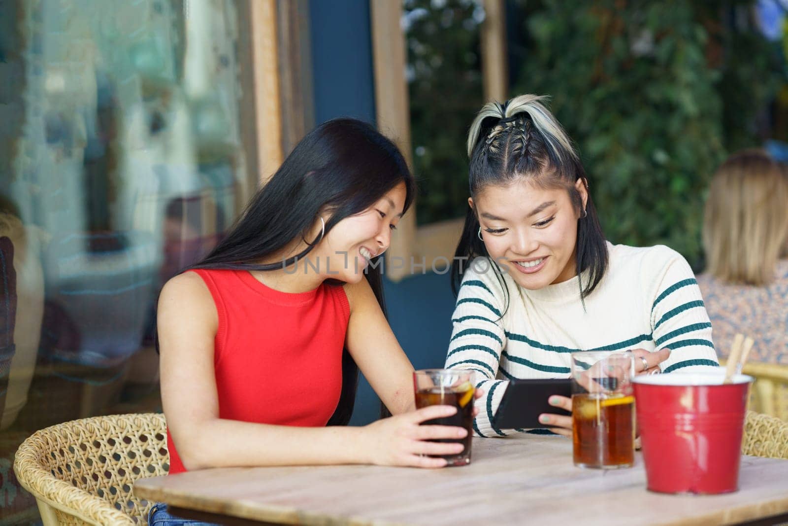 Cheerful Asian women sharing smartphone at table in cafe by javiindy