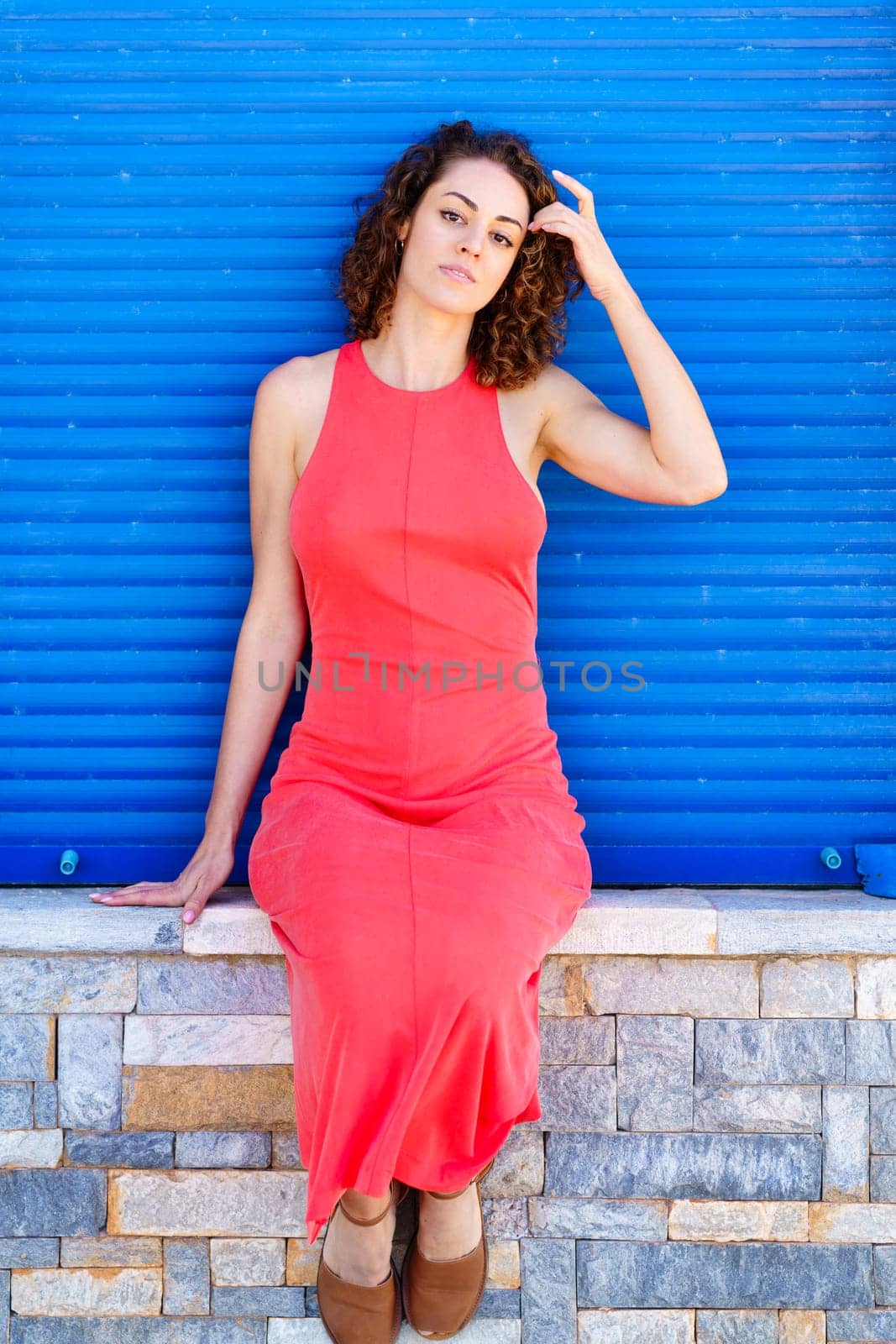 Attractive young female in red sundress, sitting on stone wall with sandals and looking at camera while touching hair with hand in daylight against blue shutters