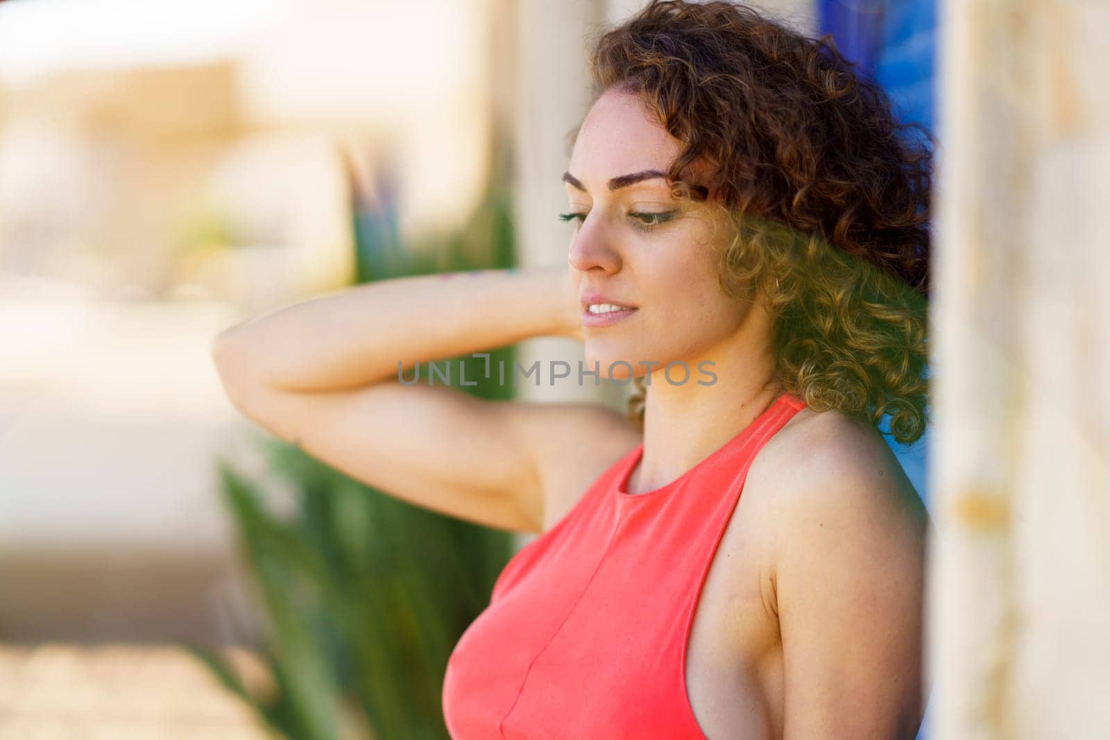 Side view of pensive young woman in sundress, with hand touching back of head standing in street near wall with blue shutters and looking down against blurred urban background