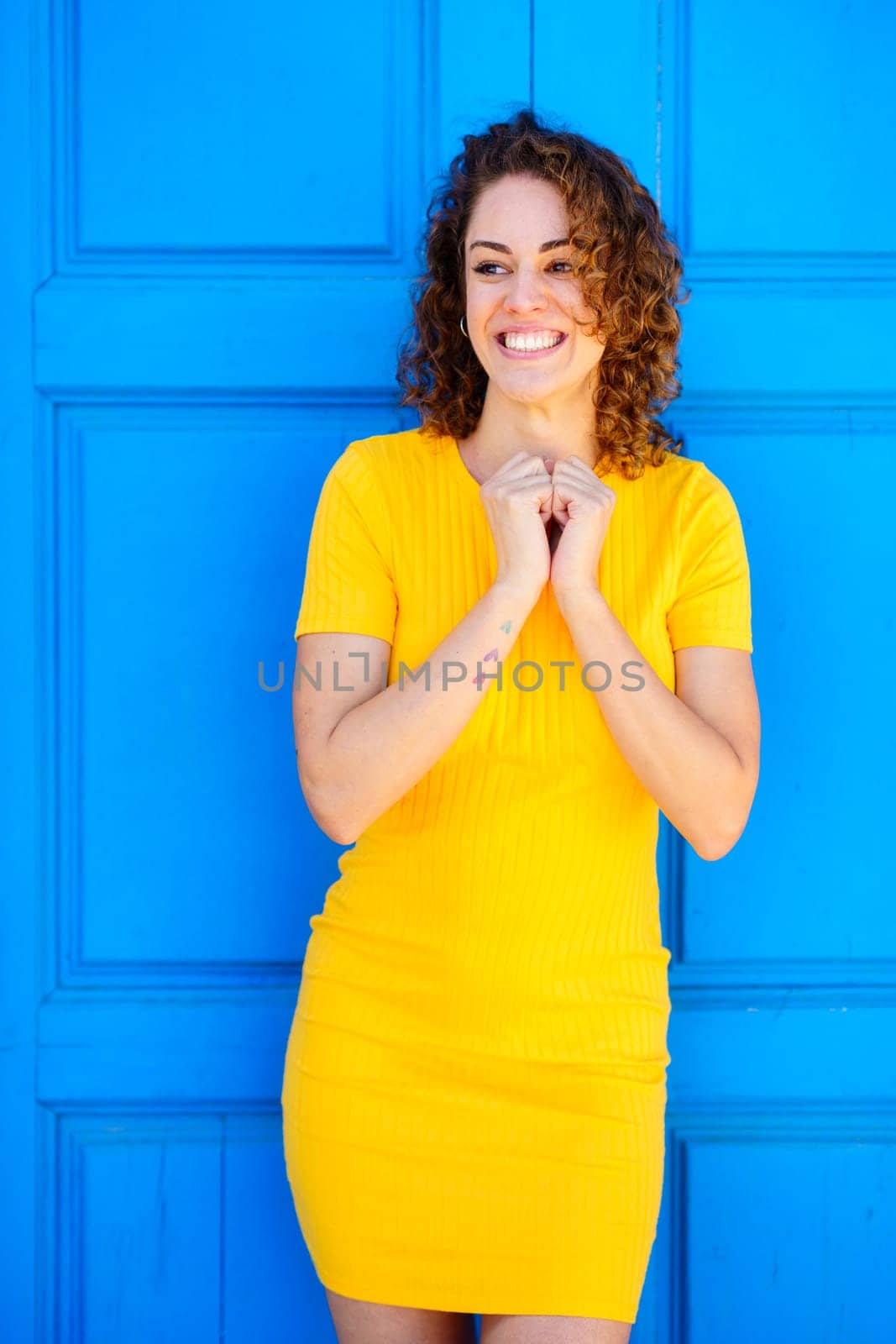 Cheerful young female in yellow dress with curly brown hair smiling and looking away against blue background on street