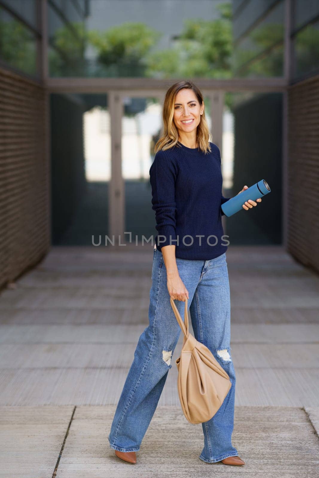 Full body happy woman, in stylish clothes with bag and thermos looking at camera with smile and walking on pavement outside modern apartment building on city street