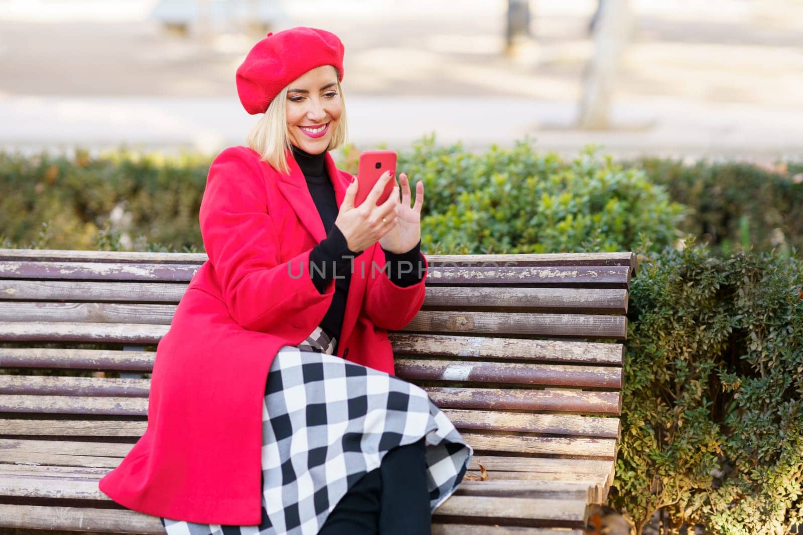 Joyful female in trendy outfit browsing modern cellphone while sitting on bench near green plants in park on autumn day