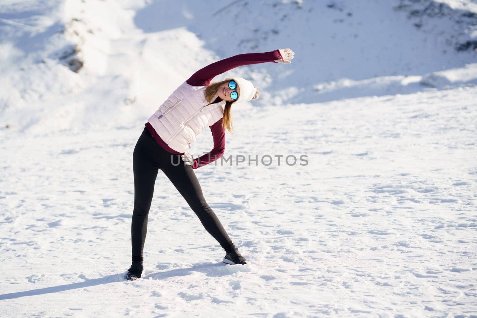 Full body of young female in sportswear and eyeglasses, stretching arm overhead to one side while practicing yoga exercise on snowy slope in winter at daylight