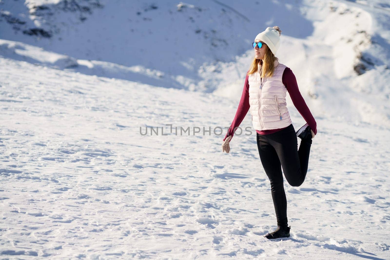 Full body of young female traveler in cozy clothes and beanie cap, standing on one leg and holding leg up while looking away on snowy slope in daylight