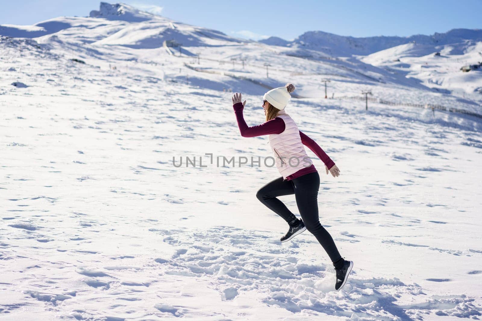 Side view of unrecognizable female traveler in warm clothes and beanie running on snowy slope of mountainous terrain in winter with blue sky