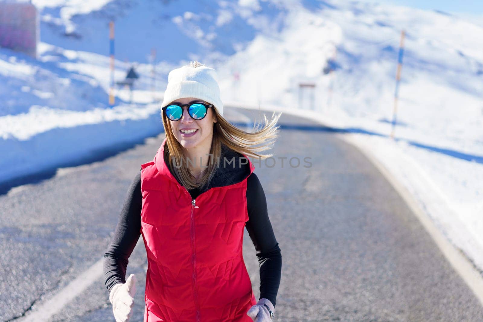 Smiling young female blonde in red vest beanie cap polarized sunglasses looking at camera, while jogging on asphalt road near snow against blurred mountain with clouds