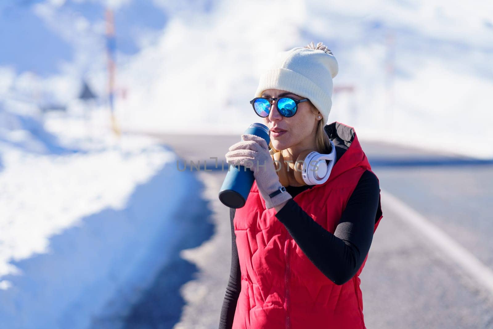 Athletic woman in outerwear and sunglasses drinking from thermos bottle having workout on road in winter mountains