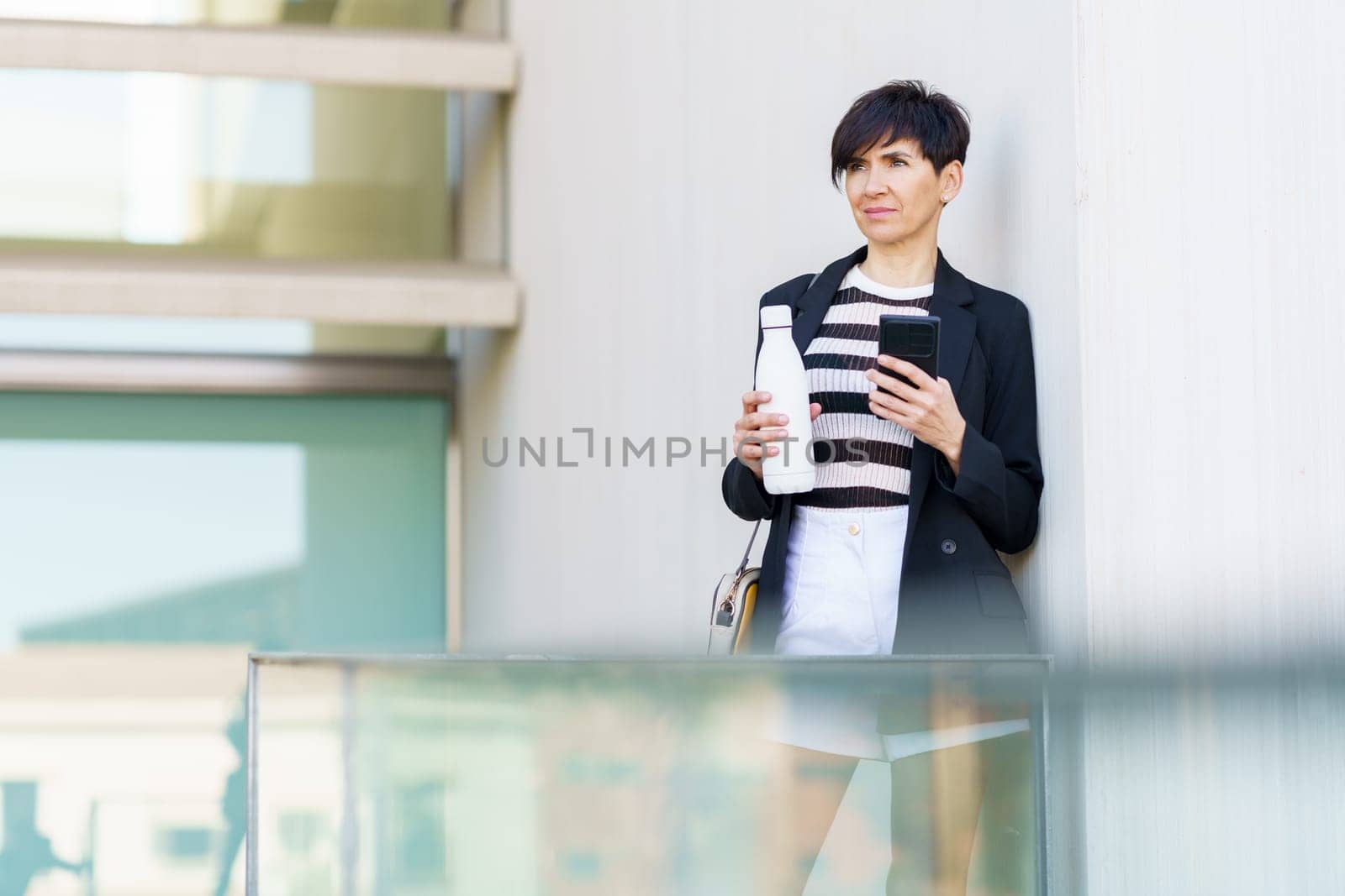 Serious businesswoman in formal clothes with handbag and smartphone leaning on hand bottle of drink while standing near glass railing