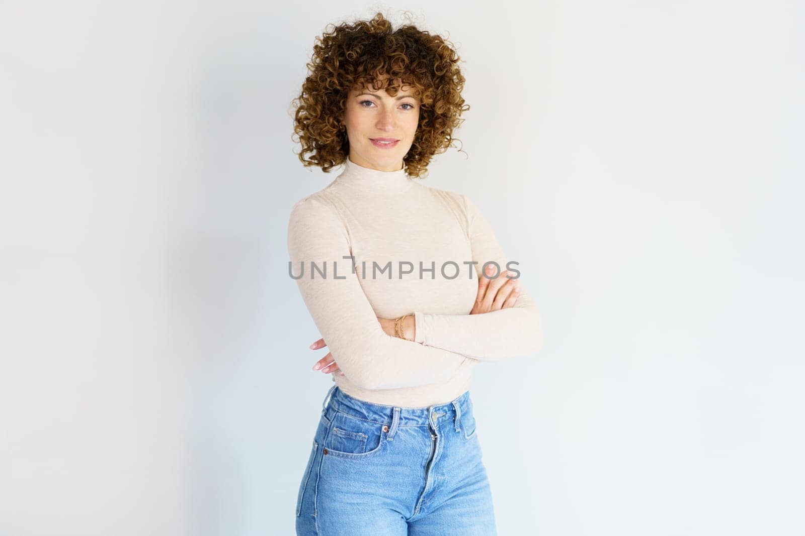 Adult curly haired female in casual clothes standing with crossed hands and looking at camera while smiling in light studio