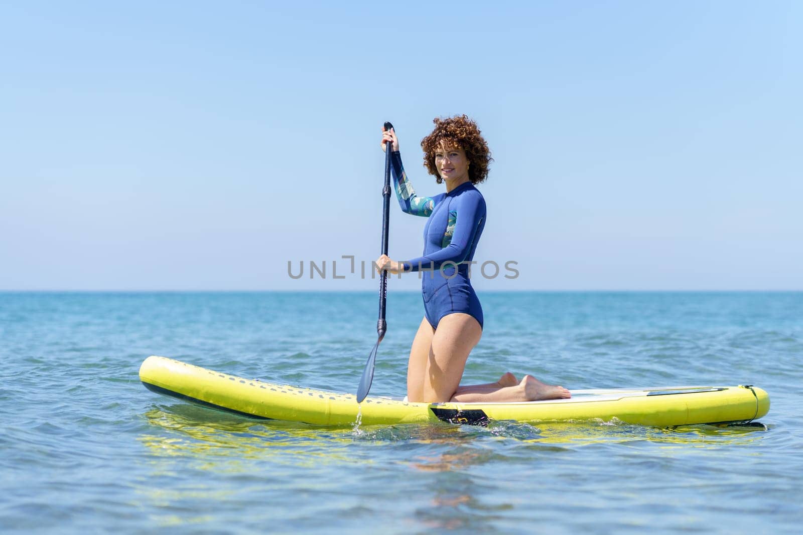 Side view of female surfer in swimsuit kneeling on paddleboard on blue sea while paddling with both hands and looking at camera in bright daylight