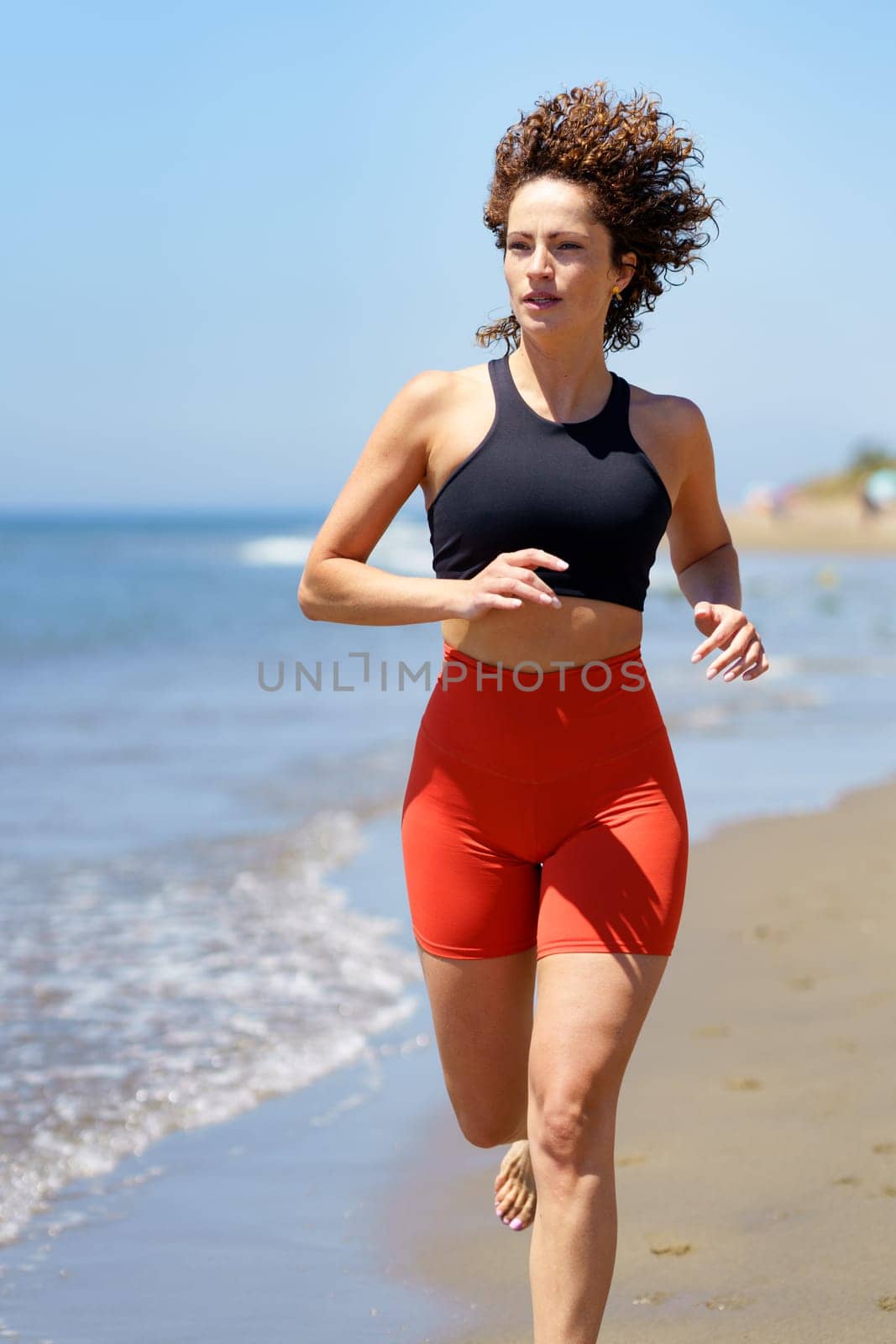 Smiling curly haired barefoot young female in sportswear warming up and looking away, while jogging alone on sandy beach near waving sea against blurred blue sky