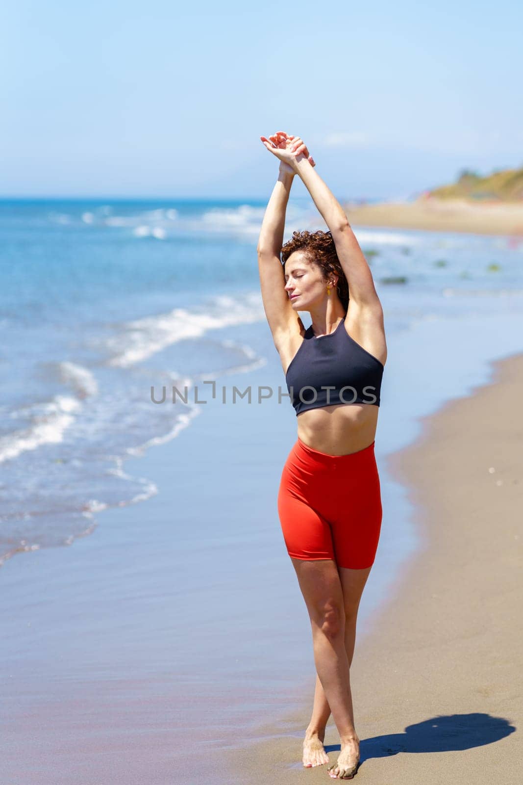 Full body of slim young sportswoman with curly hair in top and shorts standing near waving sea with raised arms while stretching body